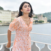 Woman in an orange and white dress standing by a waterfront with buildings and greenery in the background.